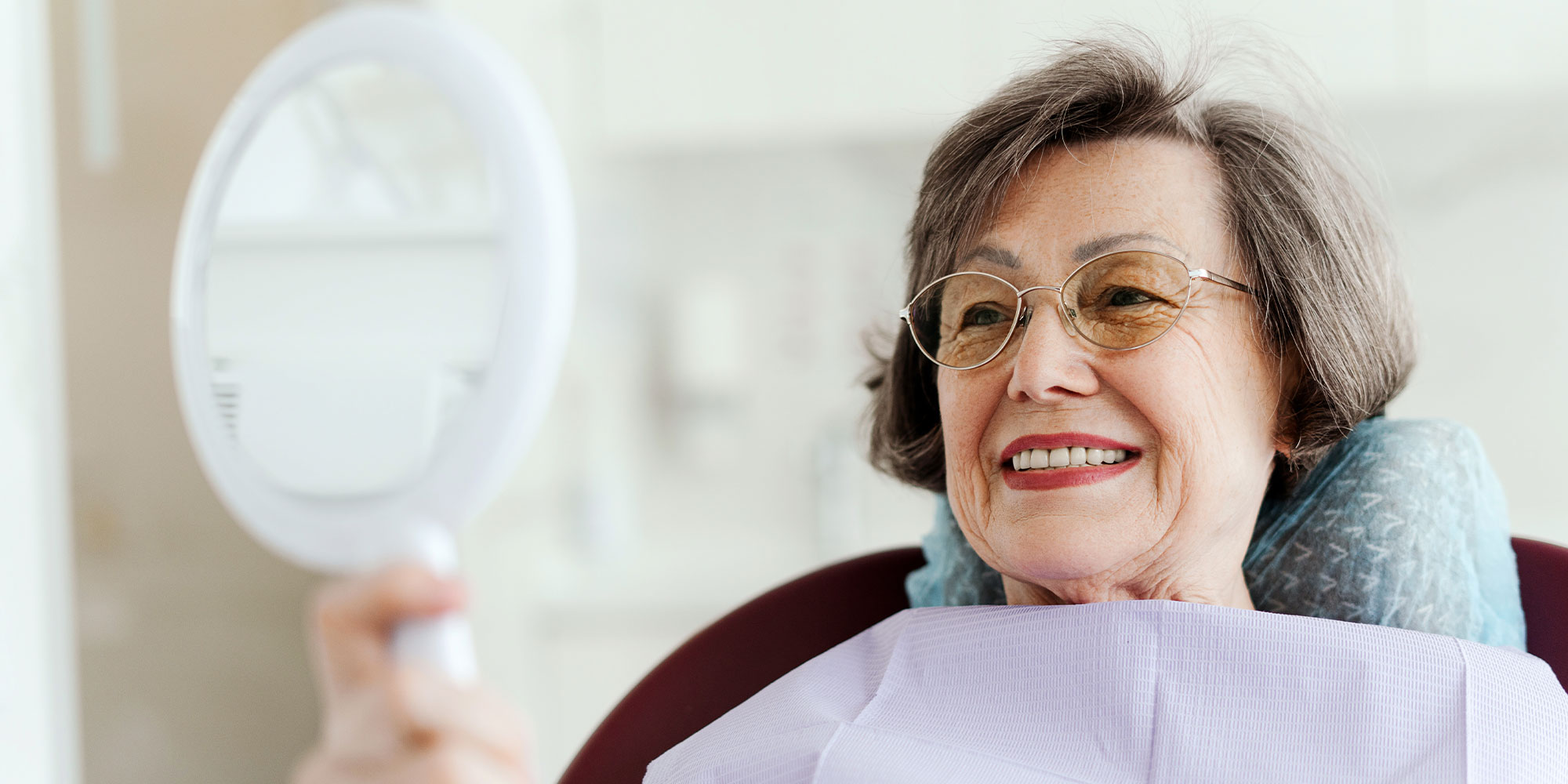 Smiling senior female patient holding mirror looking at teeth while sitting in dental chair